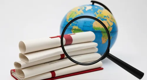 Une loupe focalisée au-dessus d'une pile de diplômes avec un globe en toile de fond, symbolisant l'examen et la validation des qualifications étrangères.