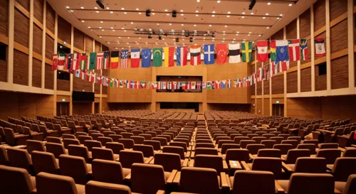 Un grand auditorium rempli de drapeaux et de bannières colorés, symbolisant diverses cultures éducatives, des chaises vides disposées pour les participants, témoignant d'une atmosphère d'anticipation et de collaboration mondiale.