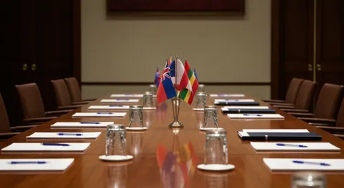 A conference room with a polished wooden table set for discussion, adorned with various country flags, notepads, and empty glasses, reflecting a serious and professional atmosphere.