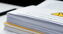 Eine Nahaufnahme eines Stapels offizieller Dokumente mit einem Warnsymbol, das die Ernsthaftigkeit der Bildungspolitik unterstreicht.